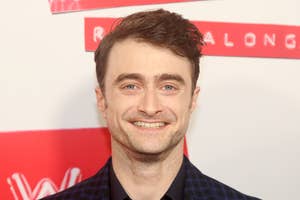 Daniel Radcliffe smiling at camera, wearing a dark plaid suit jacket over a blue shirt. Red event backdrop with logos