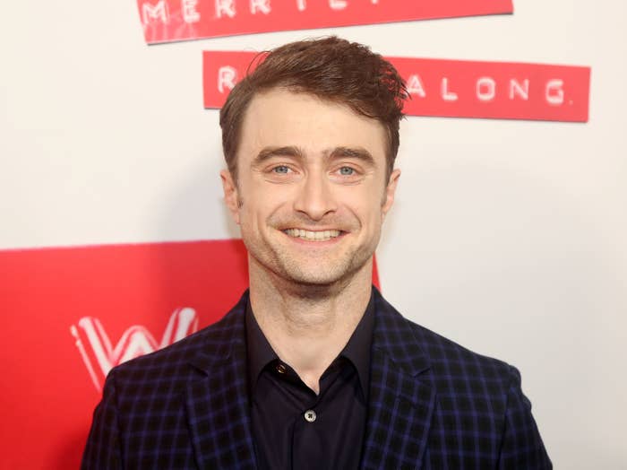 Daniel Radcliffe smiling at camera, wearing a dark plaid suit jacket over a blue shirt. Red event backdrop with logos