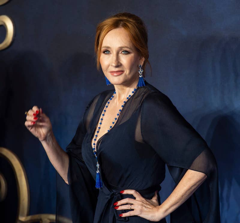 J.K. Rowling posing at an event, wearing a dark v-neck dress with blue necklace and earrings