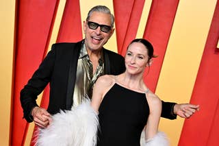 Jeff Goldblum in a patterned shirt and glasses; with a woman in a feathered outfit on a red carpet