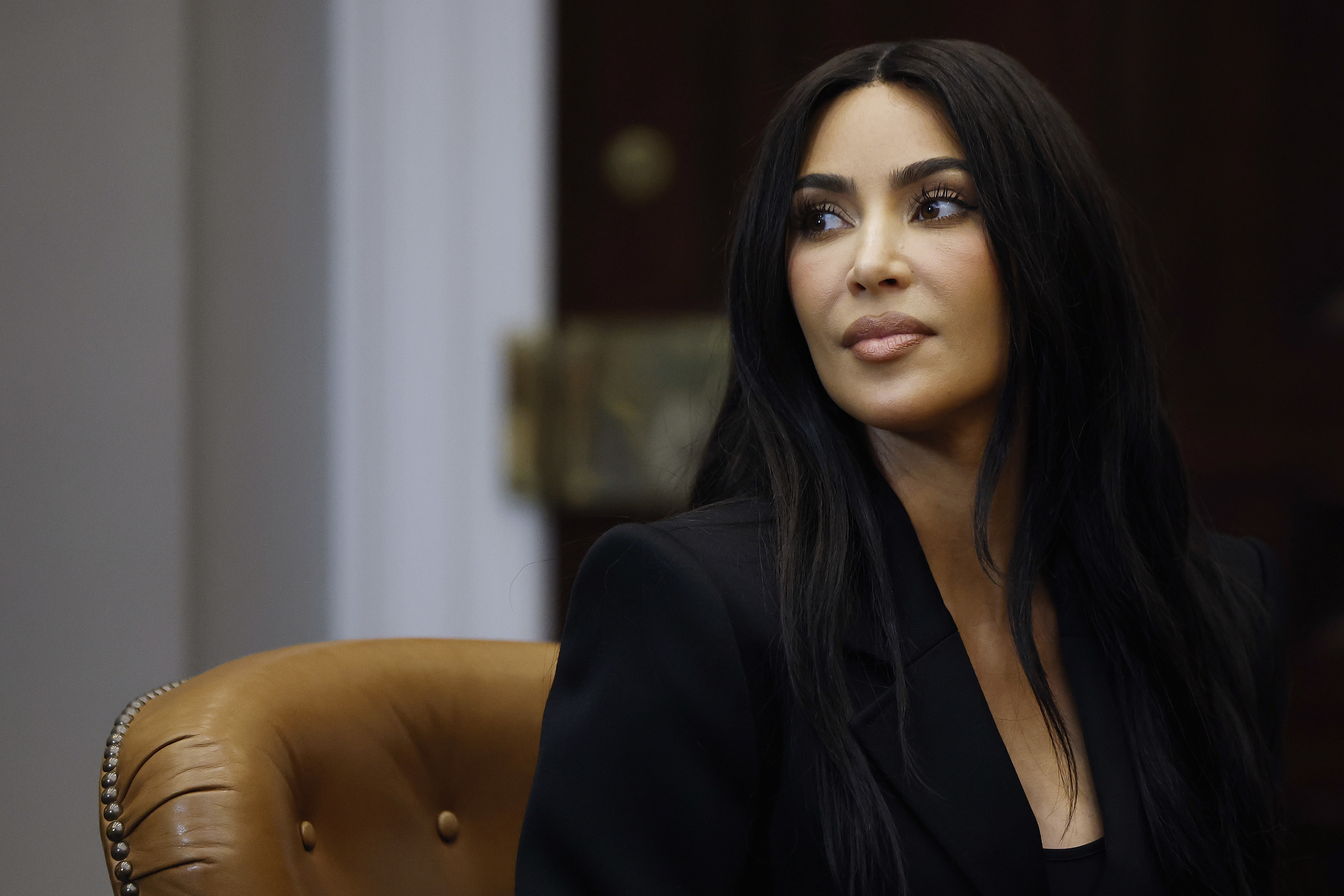 Kim Kardashian in a black blazer, seated, looking to the side with a focused expression