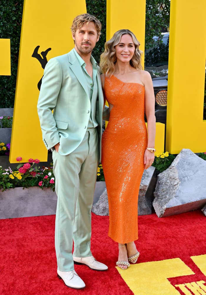 Two individuals posing on the red carpet; one in a light-toned suit and the other in a one-shoulder sequined gown