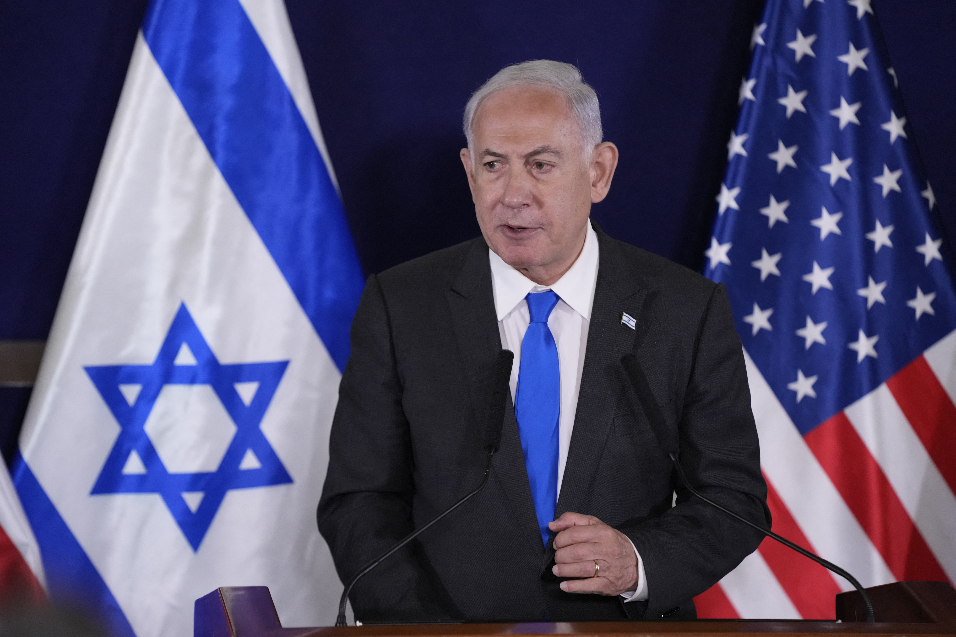 Benjamin Netanyahu stands at a podium with Israeli and American flags in the background. He is wearing a dark suit