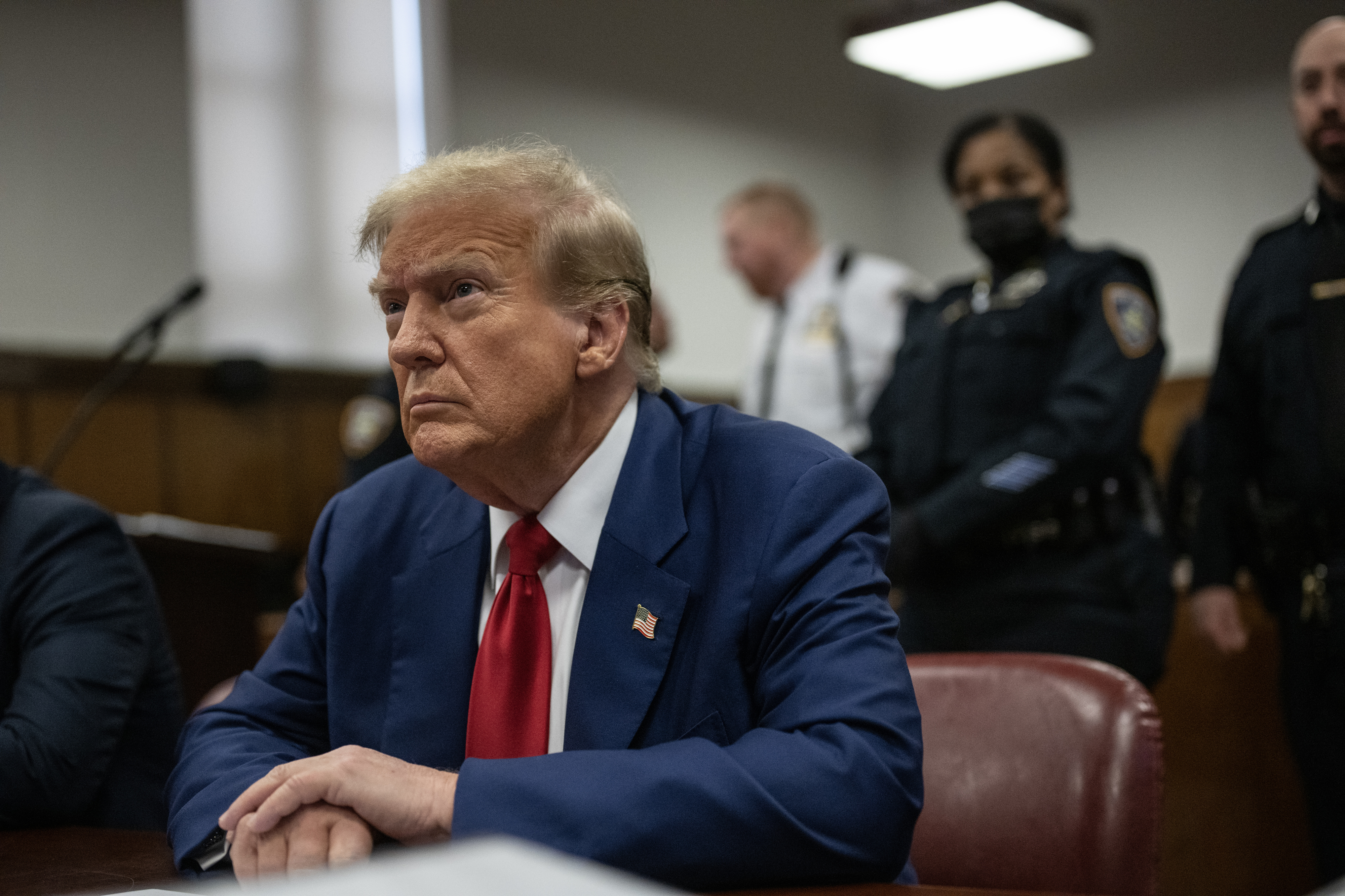Donald Trump seated in a courtroom with security personnel standing behind him