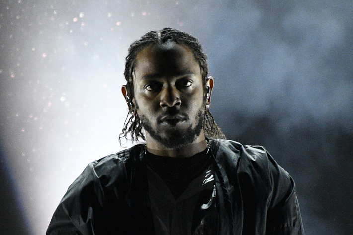 Kendrick Lamar performs on stage, wearing a black jacket, intense expression with a dark, moody backdrop