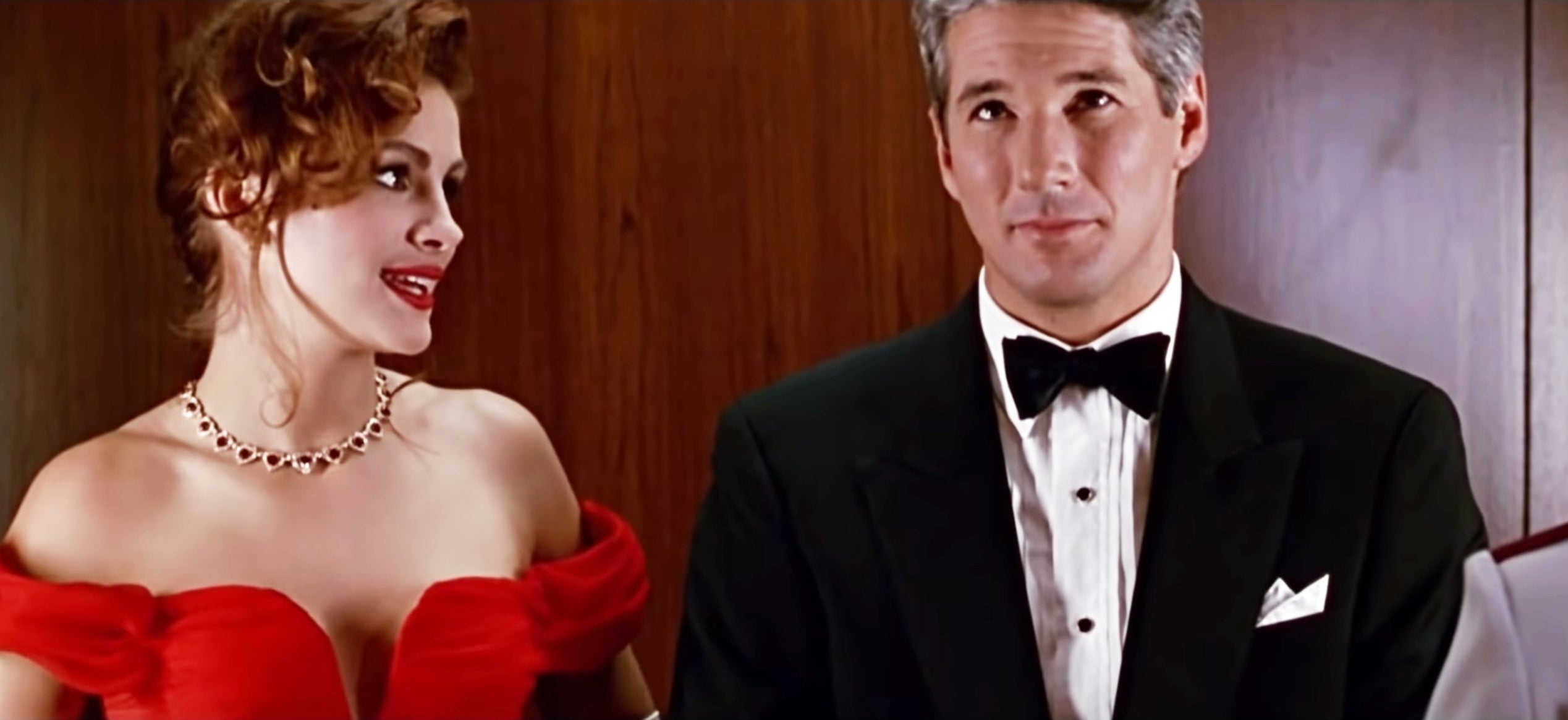 Two actors in formal attire; a woman in a red off-shoulder dress and a man in a black tuxedo, standing side by side