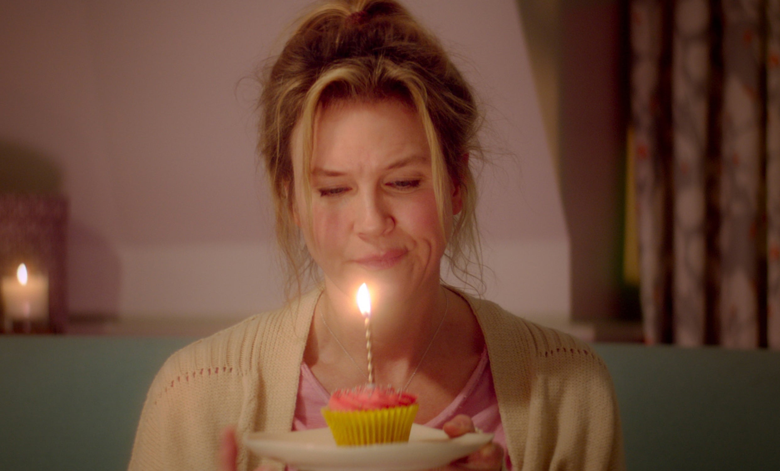 Renee Zellweger in a scene, holding a cupcake with a lit candle, appearing contemplative