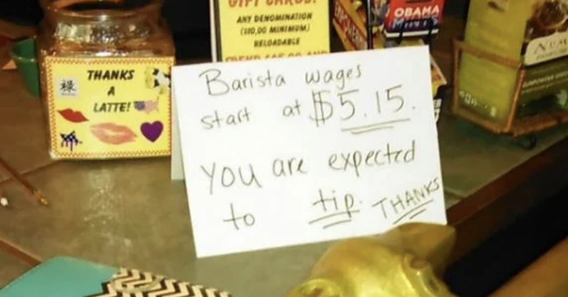 Handwritten sign states &quot;Barista wages start at $5.15. You are expected to tip. Thanks&quot; on a cafe counter
