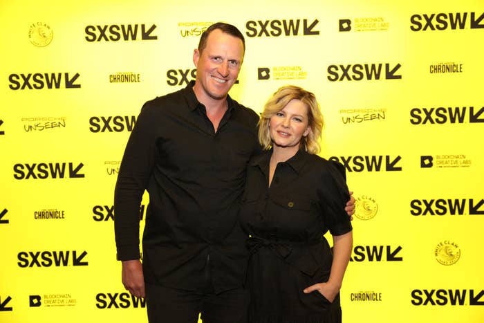 Two people standing together against a backdrop with &#x27;SXSW&#x27; logos; both are dressed in black, one in a buttoned shirt, the other in a dress