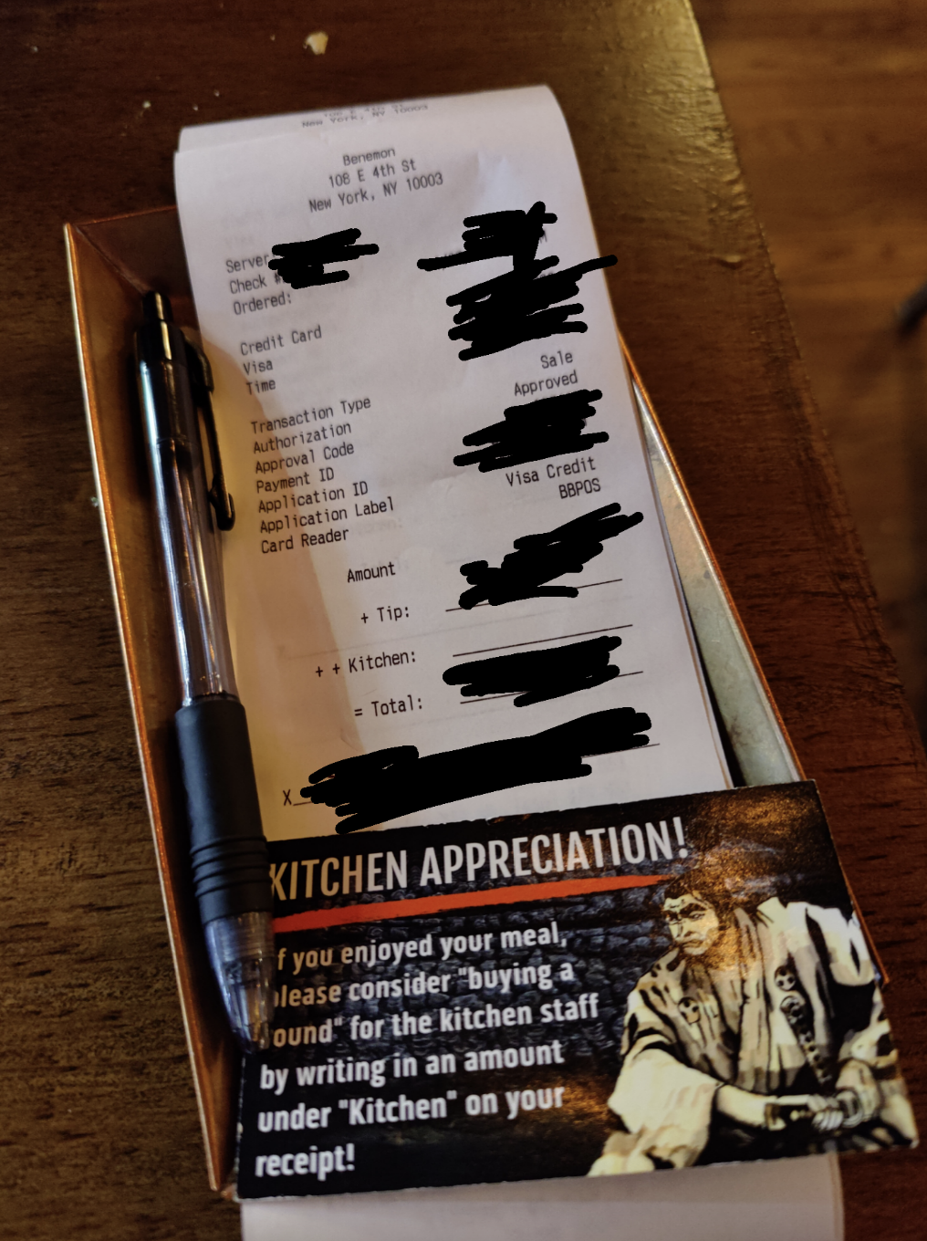 Restaurant receipt with handwritten tip and total amount on wood table, pen aside, note suggesting a tip for kitchen staff
