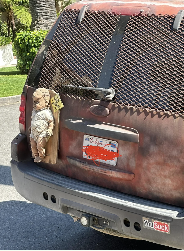 A truck&#x27;s rear window features a decal simulating a child and dog peering out. Bumper stickers and a license plate are visible