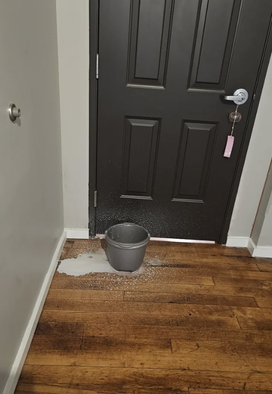 A bucket catches water drips under a leak by a closed black door indoors