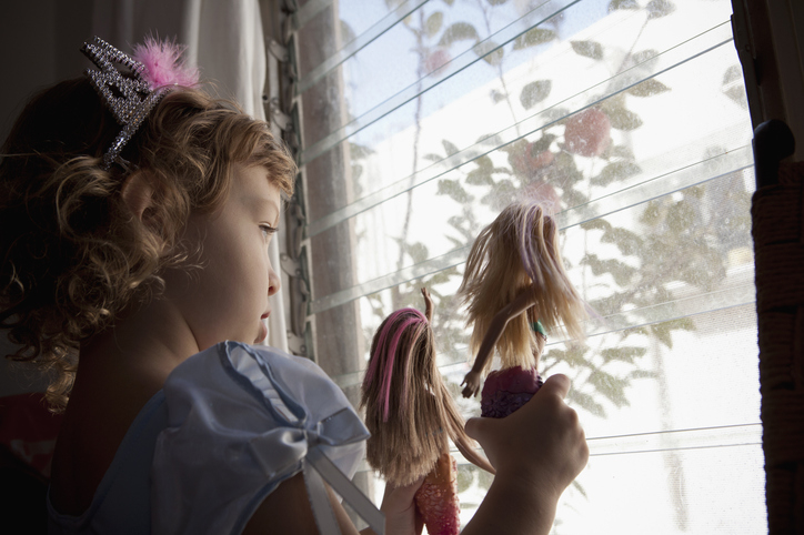 Child in tiara plays with dolls by a window, looking outside