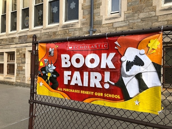 Banner for a Scholastic Book Fair displayed on a fence, with cartoon characters and a message about benefiting the school