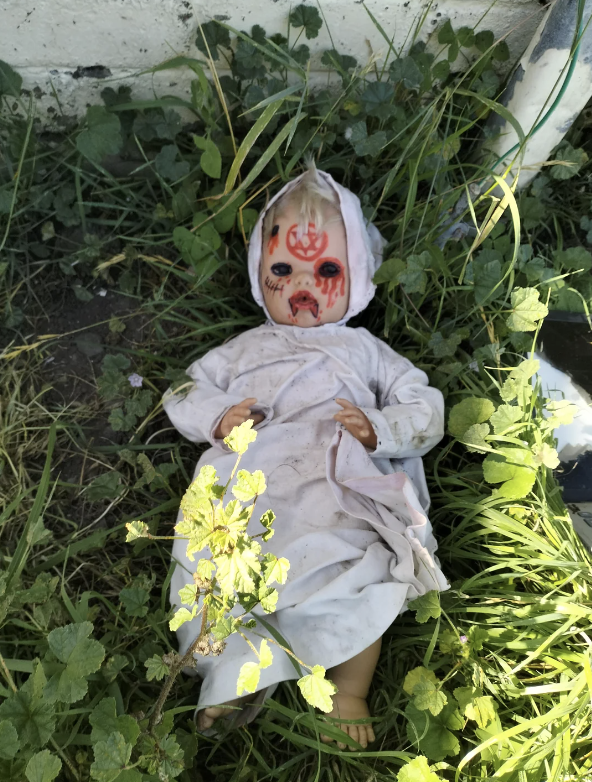 A doll with a scary face paint lying in overgrown grass