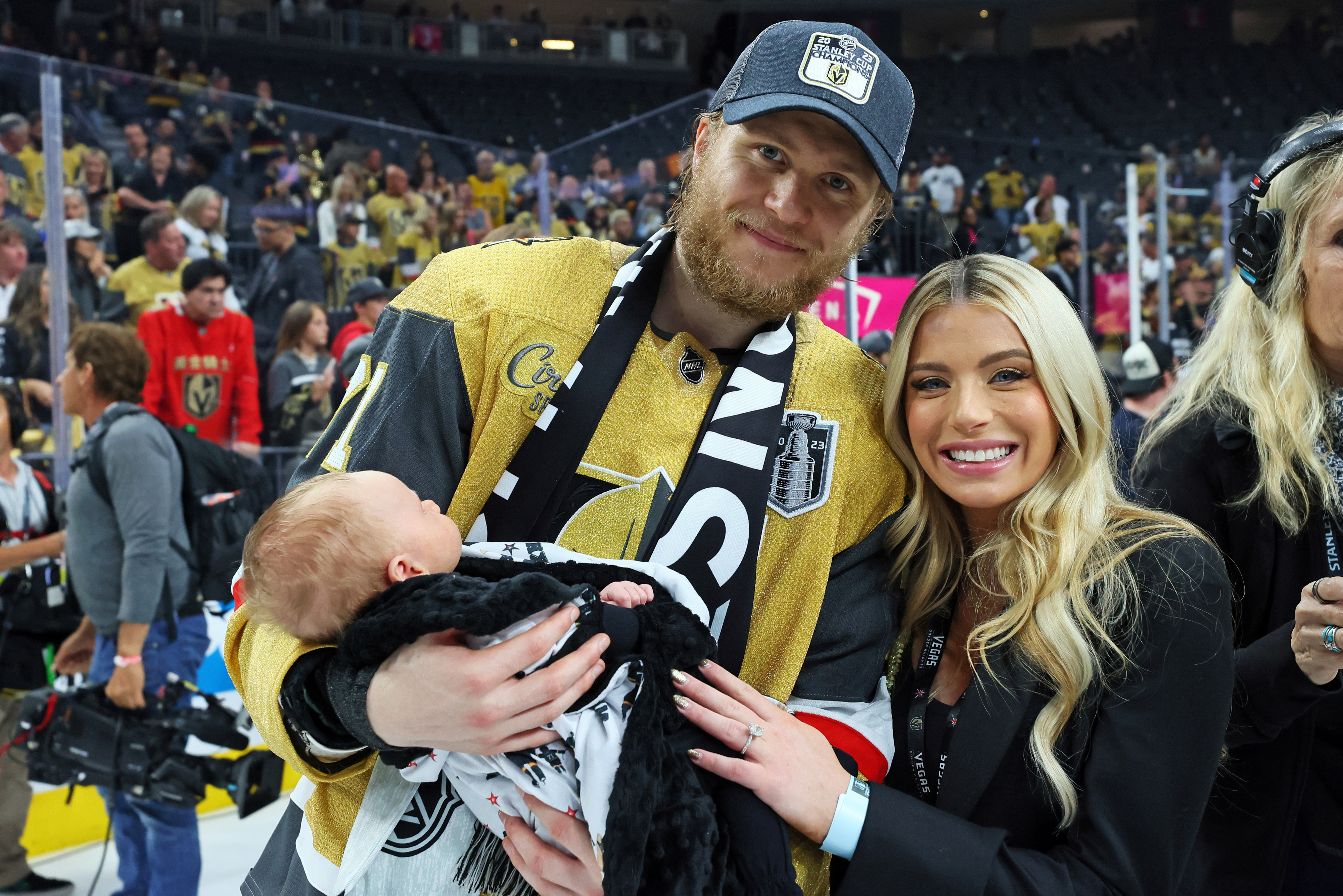 Man in hockey jersey holding a baby, standing next to a woman, at a sports event