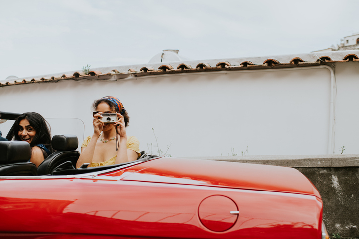 Two people smiling in a red convertible, one adjusting sunglasses, evoking a sense of carefree nostalgia