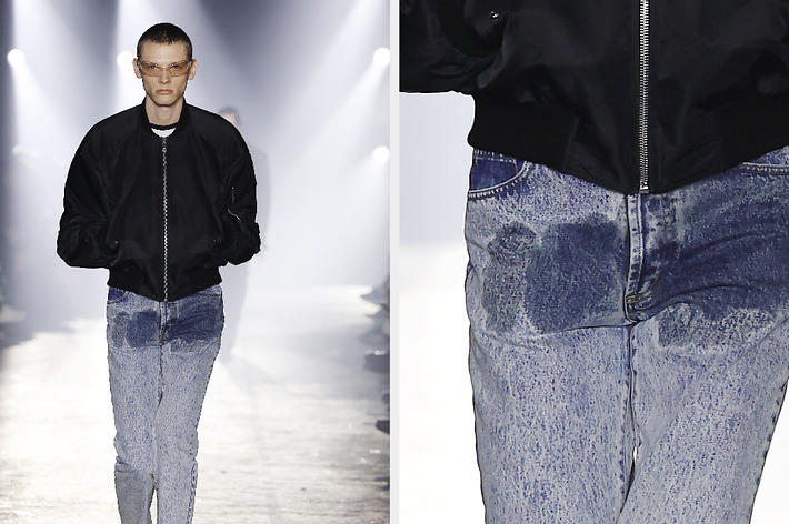 Model on runway in black jacket and faded jeans with wet stain in groin. Close-up shows focus on urine-like stain on denim.