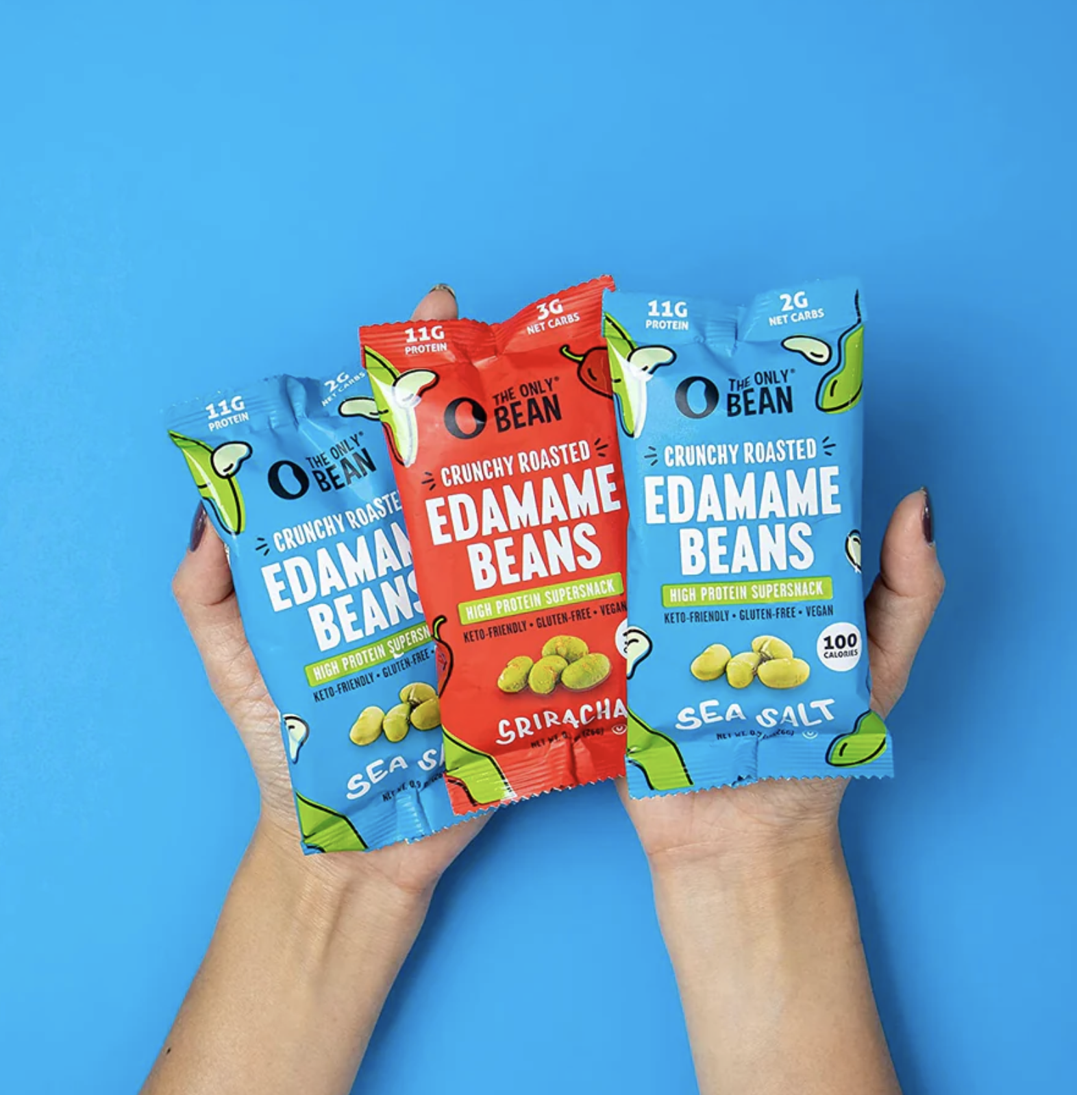 Two hands holding three different flavors of crunchy roasted edamame bean snack packs