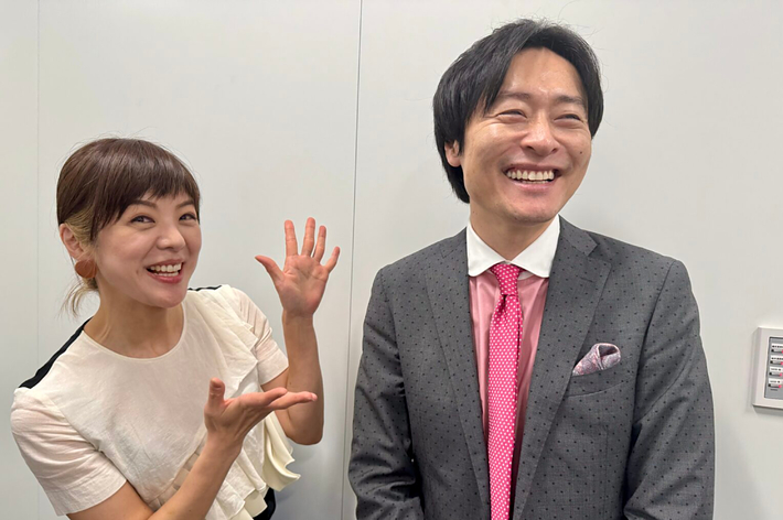 二人の笑顔の男女がカメラに向かって立っていて、男性がスーツを着用、女性はカジュアルな服を着ています。