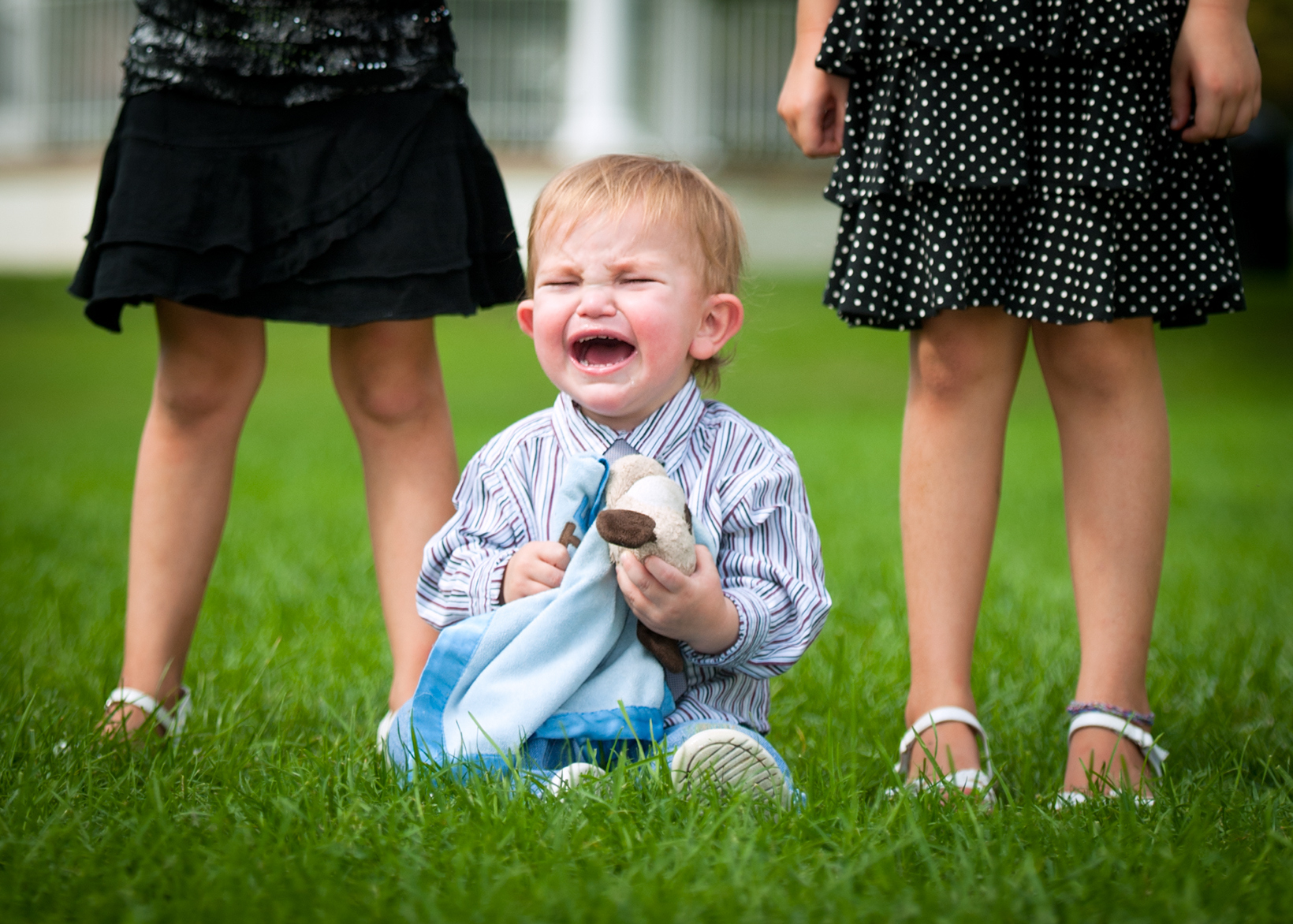 Toddler crying on grass between two people, holding a blanket and wearing a striped shirt