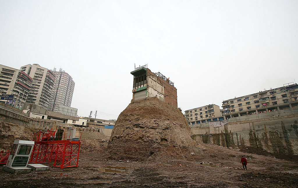 A lone building stands atop an isolated dirt mound amidst a construction site surrounded by apartment buildings