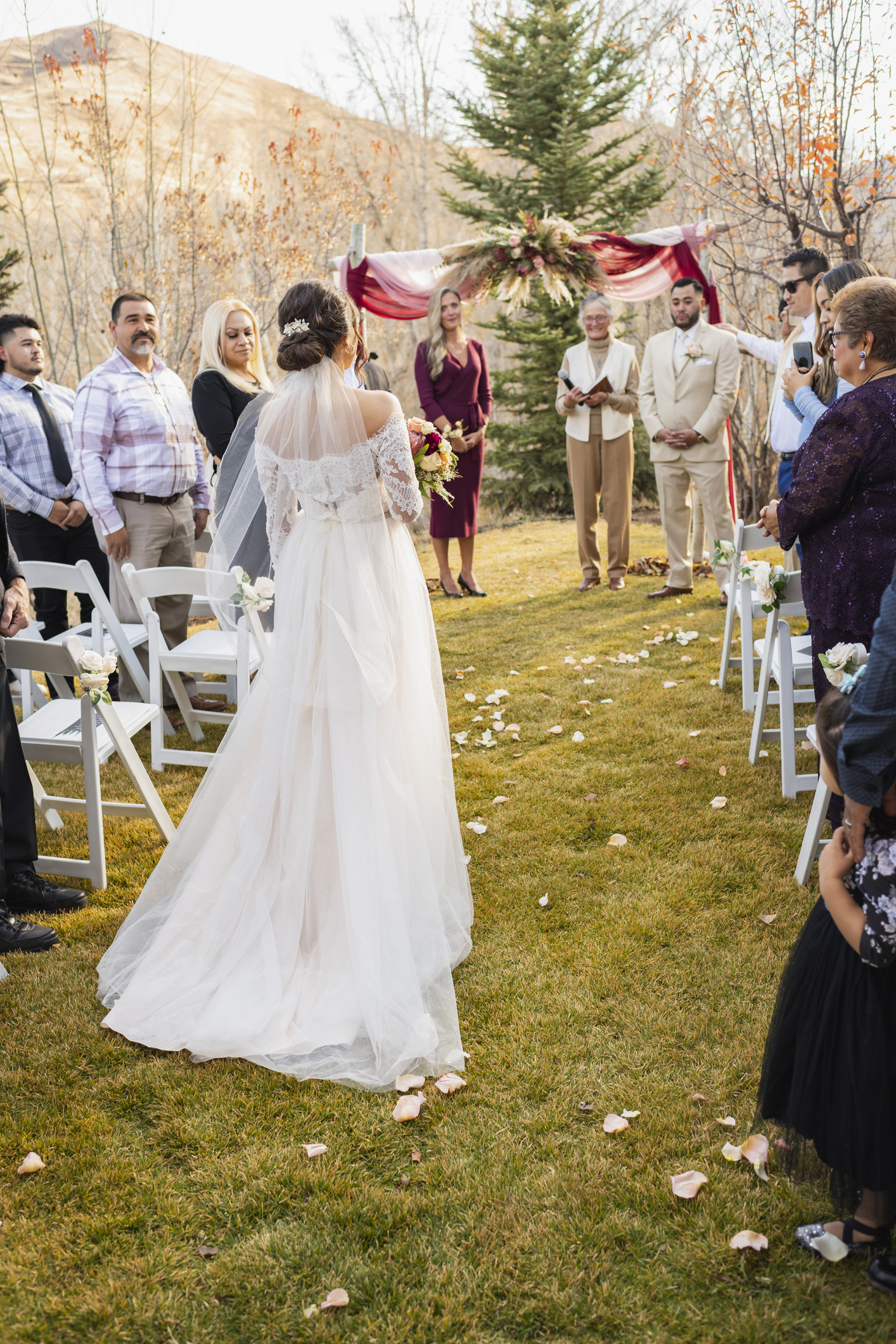 Bride in a flowing white dress holding a bouquet walks down the aisle at an outdoor wedding ceremony with guests on either side