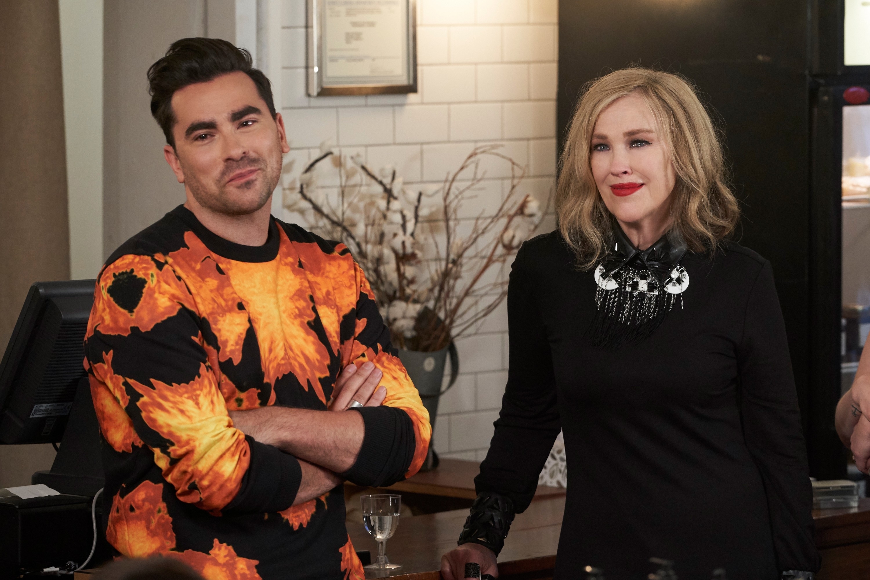 David Rose and Moira Rose from Schitt&#x27;s Creek stand side by side; he wears a flame-patterned sweater, she in a black dress with a statement necklace