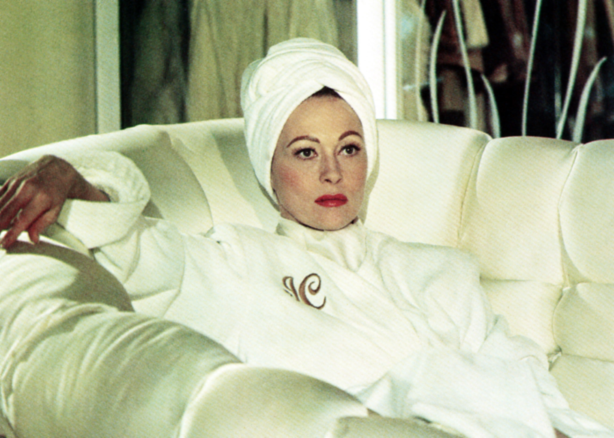 Faye Dunaway in a white outfit with a head wrap sits on a white couch, posing with a sophisticated demeanor