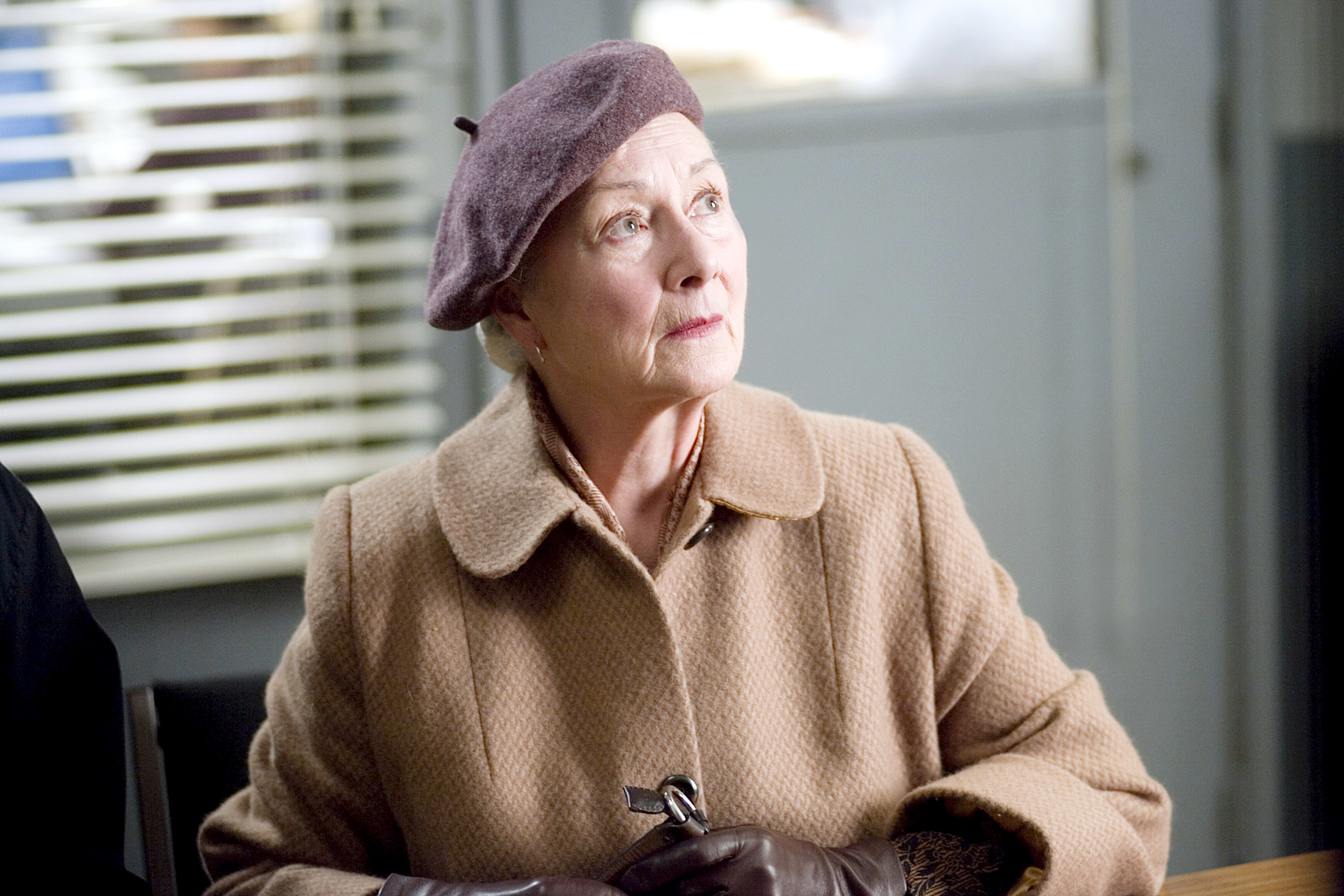 Rosemary Harris in a tan coat and purple hat looks upwards with a pensive expression
