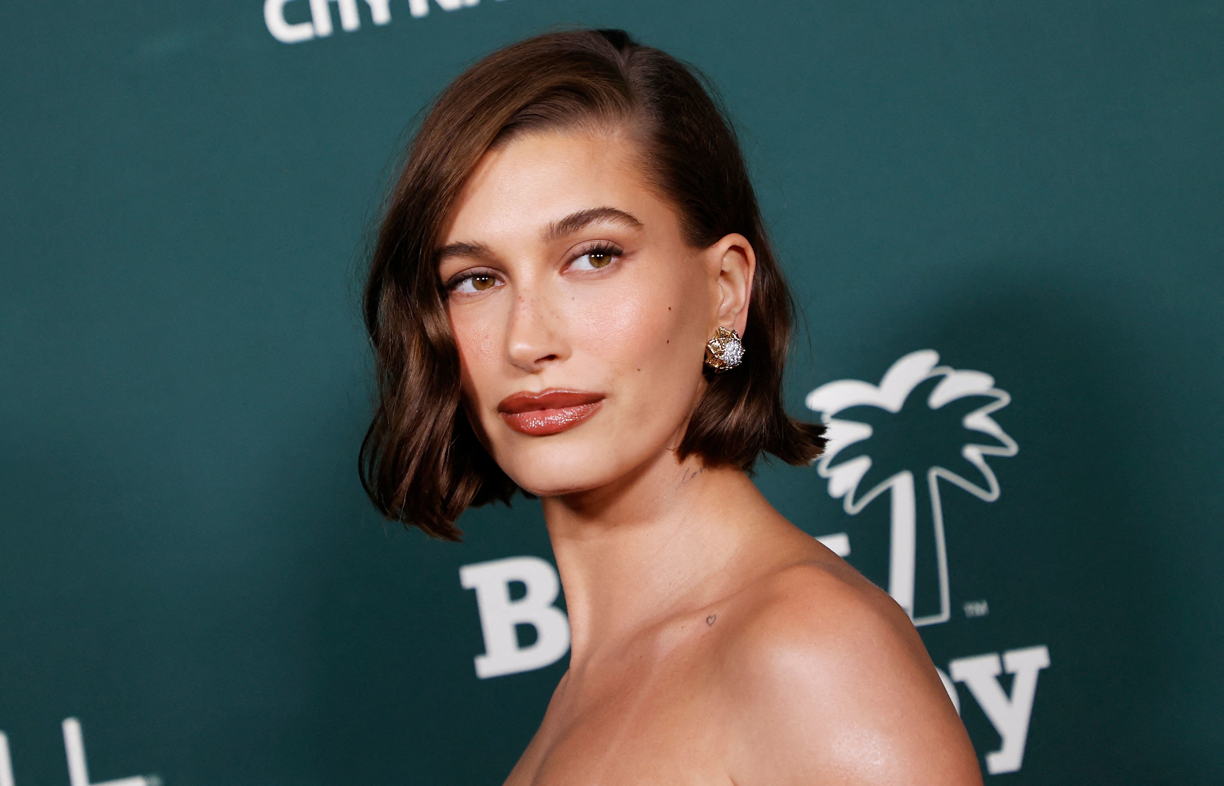 Hailey Bieber poses at an event wearing a chic shoulder-baring outfit with statement earrings