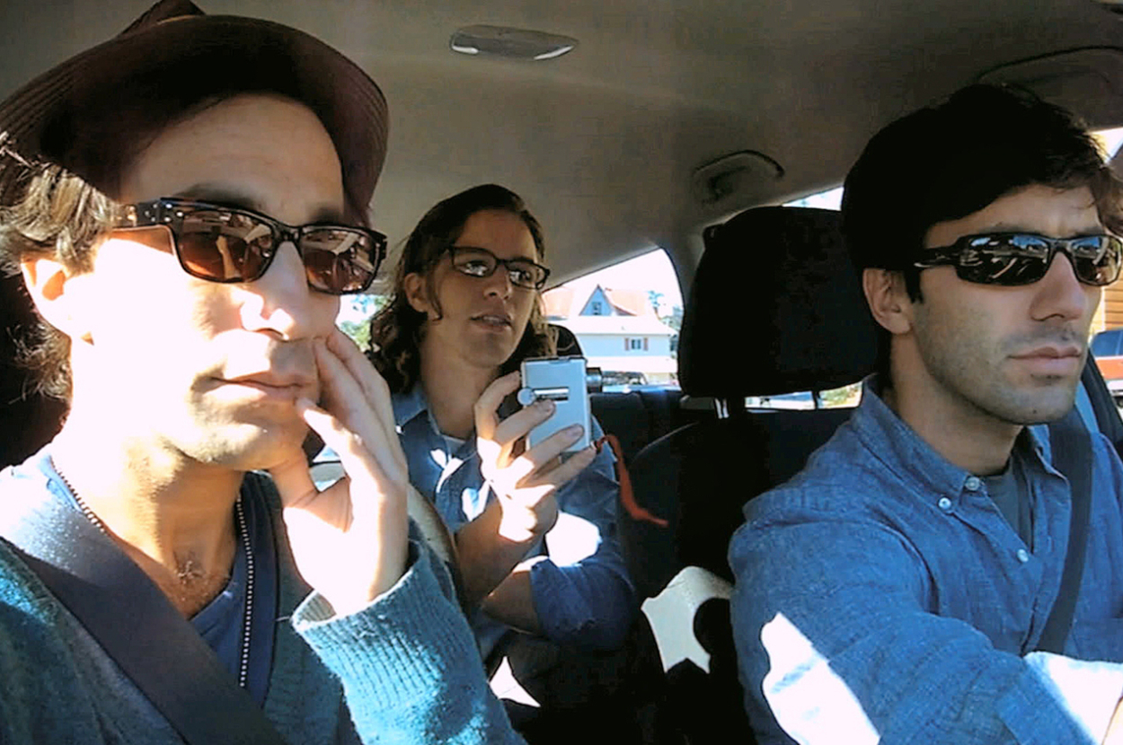 Three people in a car; one in front passenger seat looking forward, driver glancing aside, person in back using a smartphone