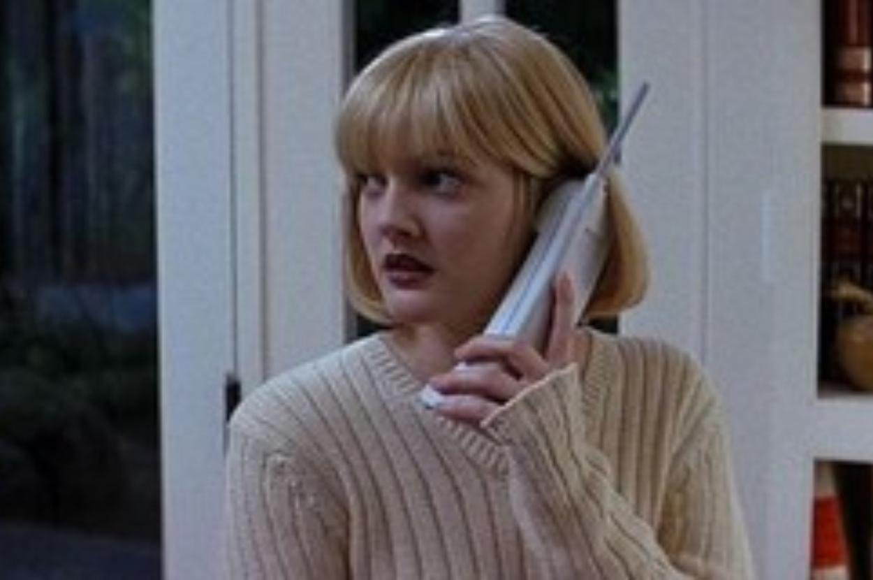 Drew Barrymore as Casey Becker in "Scream," holding a cordless phone, looking worried. She wears a knitted sweater