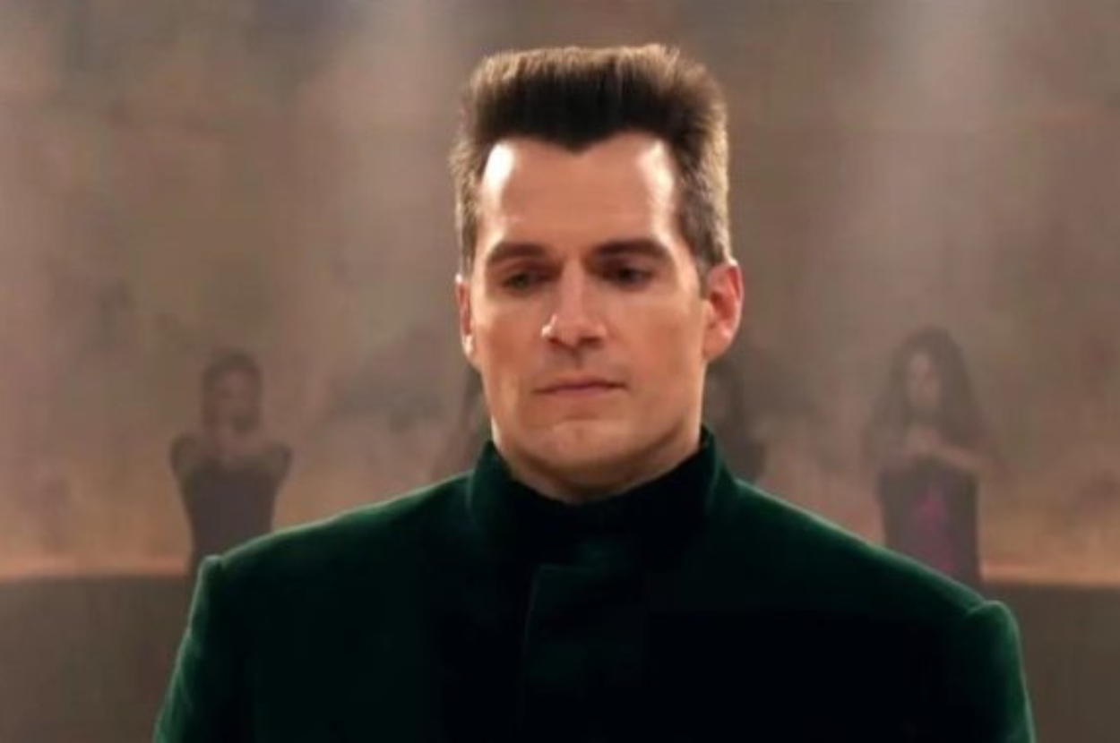 A man with dark hair wearing a green high-neck outfit stands in front of a blurred background
