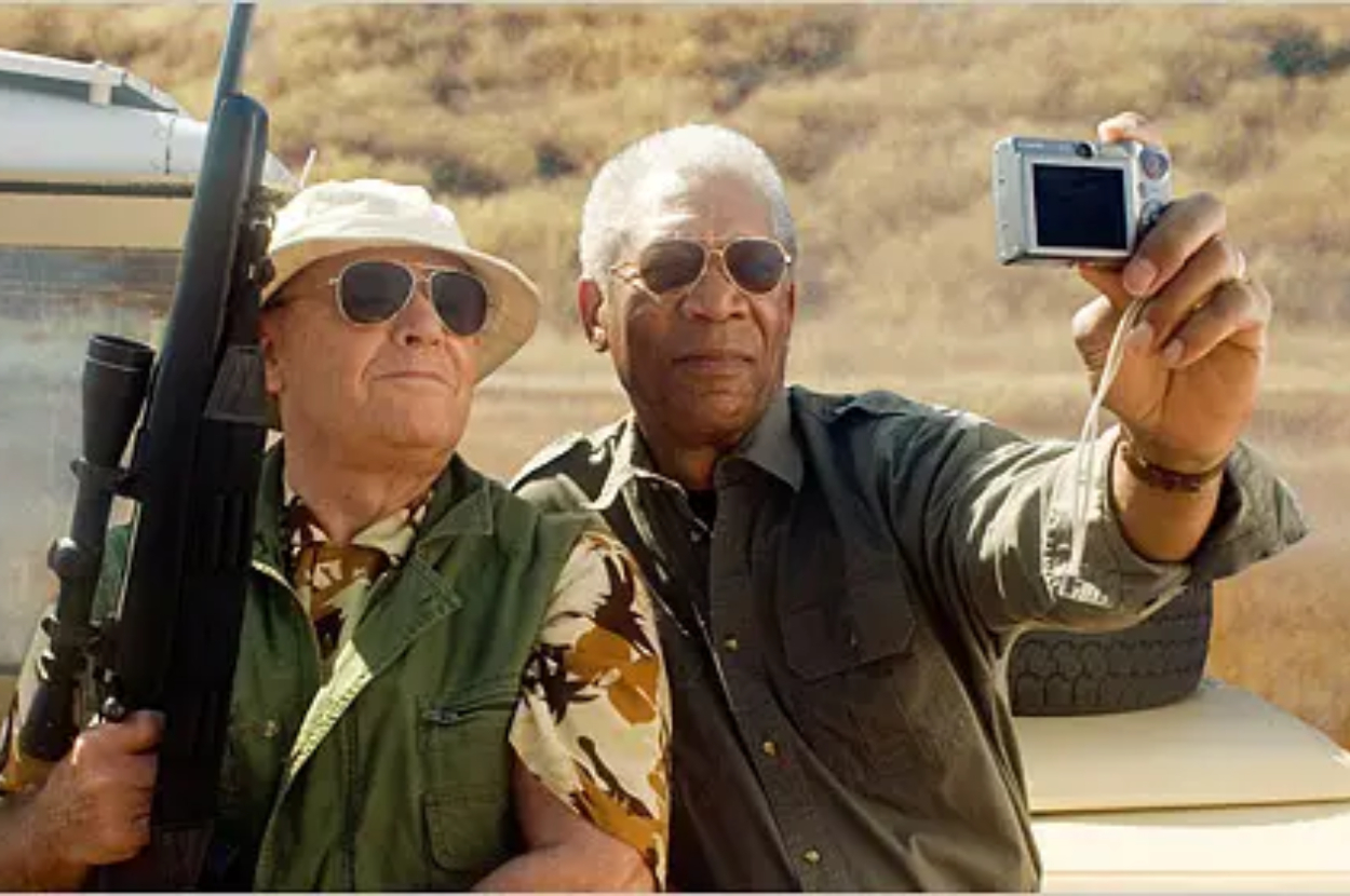 Two men, one holding a camera and the other a rifle, pose for a selfie with smiles. They wear casual outdoor gear