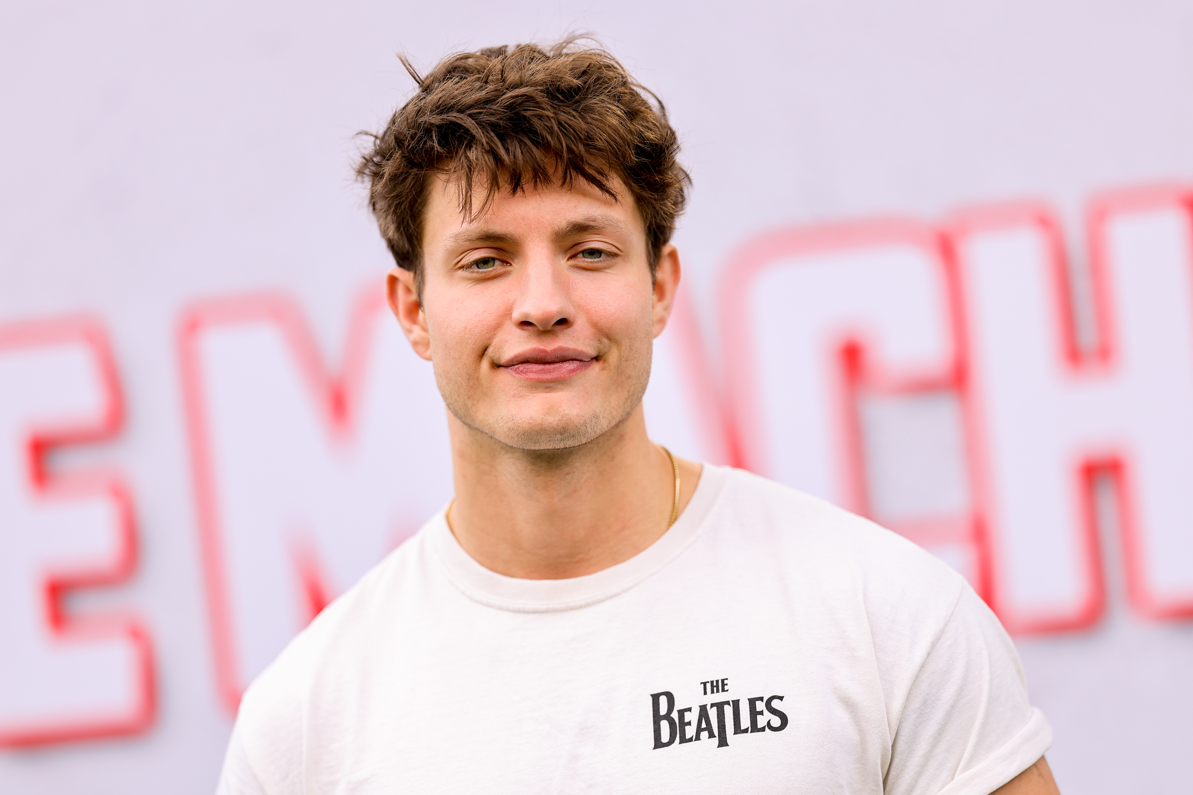 Matt Rife smiling with short hair, wearing a T-shirt with &quot;The Beatles&quot; text, in front of a blurred backdrop