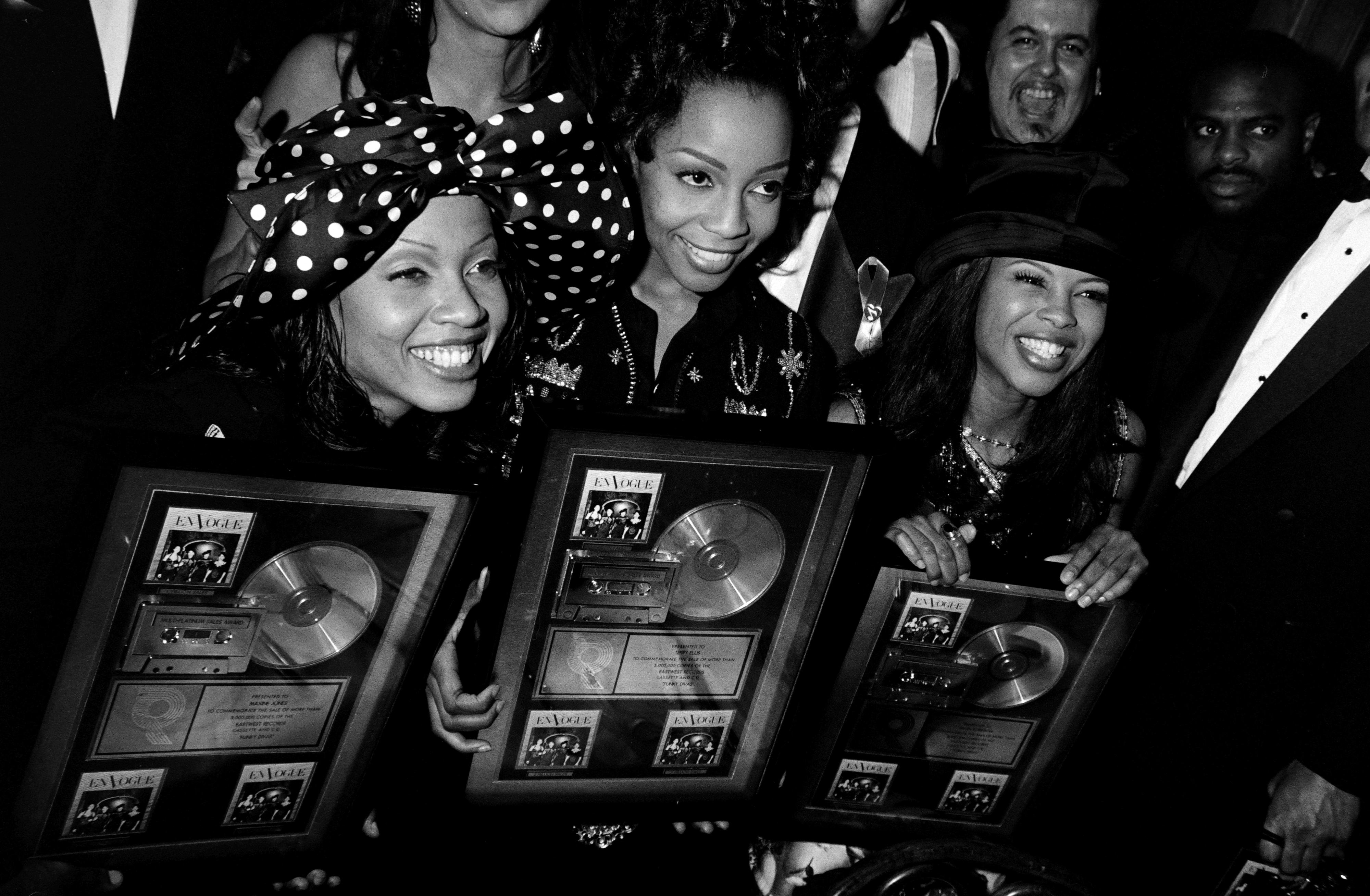 Three women smiling, holding music awards plaques, dressed in formal attire