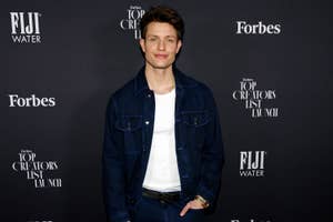 Matt Rife posing at an event wearing a denim jacket and a white shirt, standing before a backdrop with logos