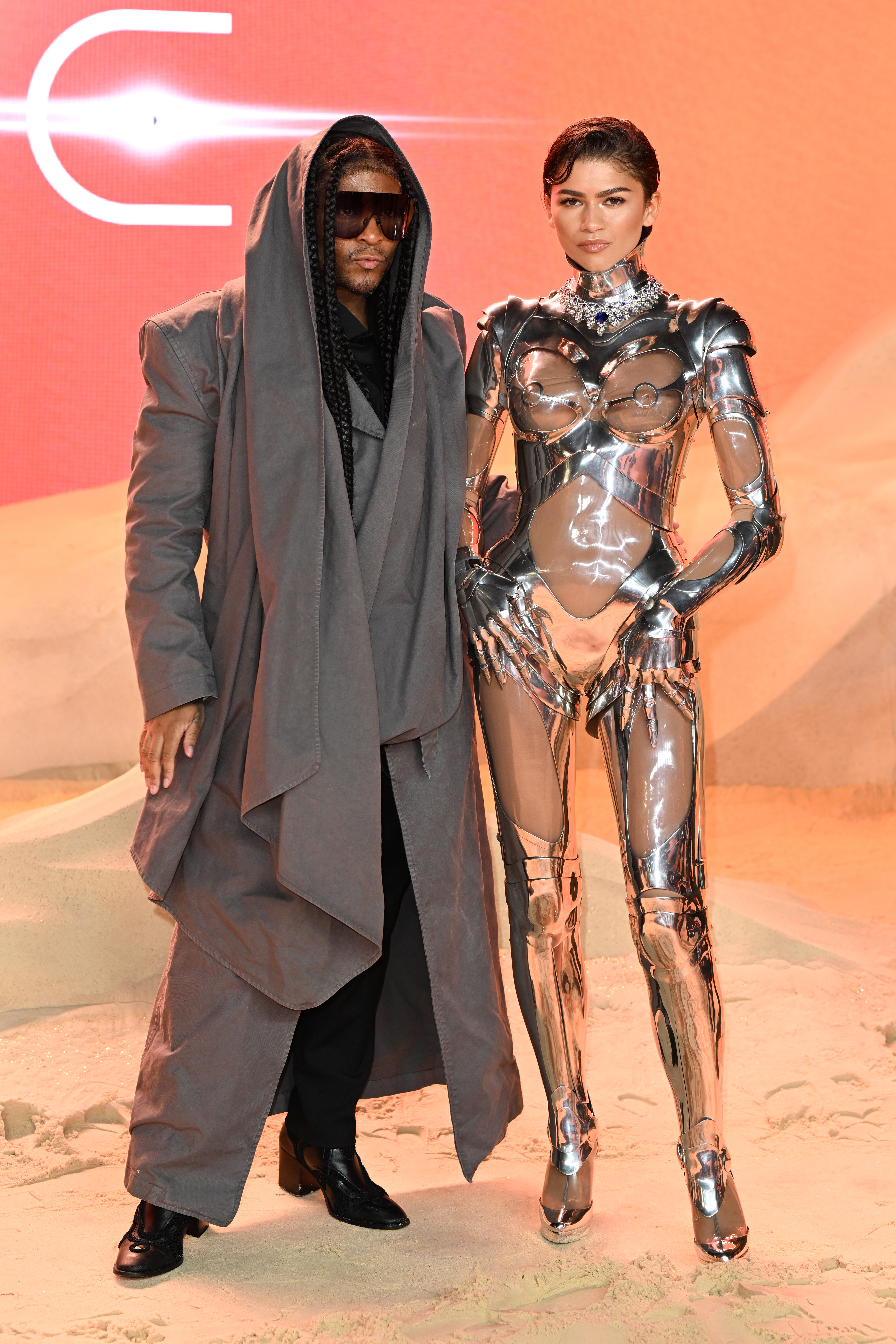 Two individuals in unique fashion attire posing together; one in a draped, hooded ensemble, and the other in a metallic bodysuit