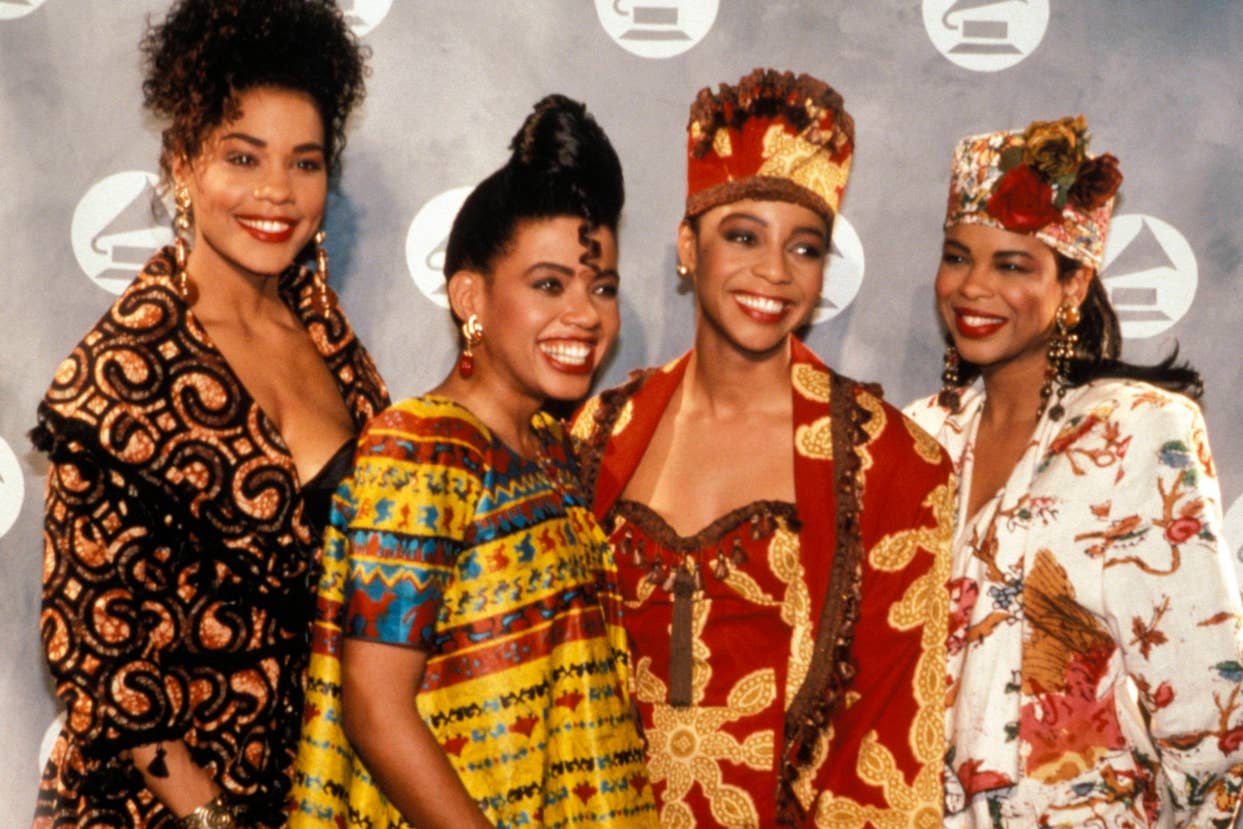 Four women in unique, patterned attire pose together. The second from the left wears a traditional headpiece