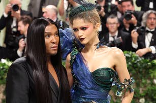 Two individuals on the red carpet, one in a sleek black outfit, another in a detailed blue gown with an avant-garde headpiece