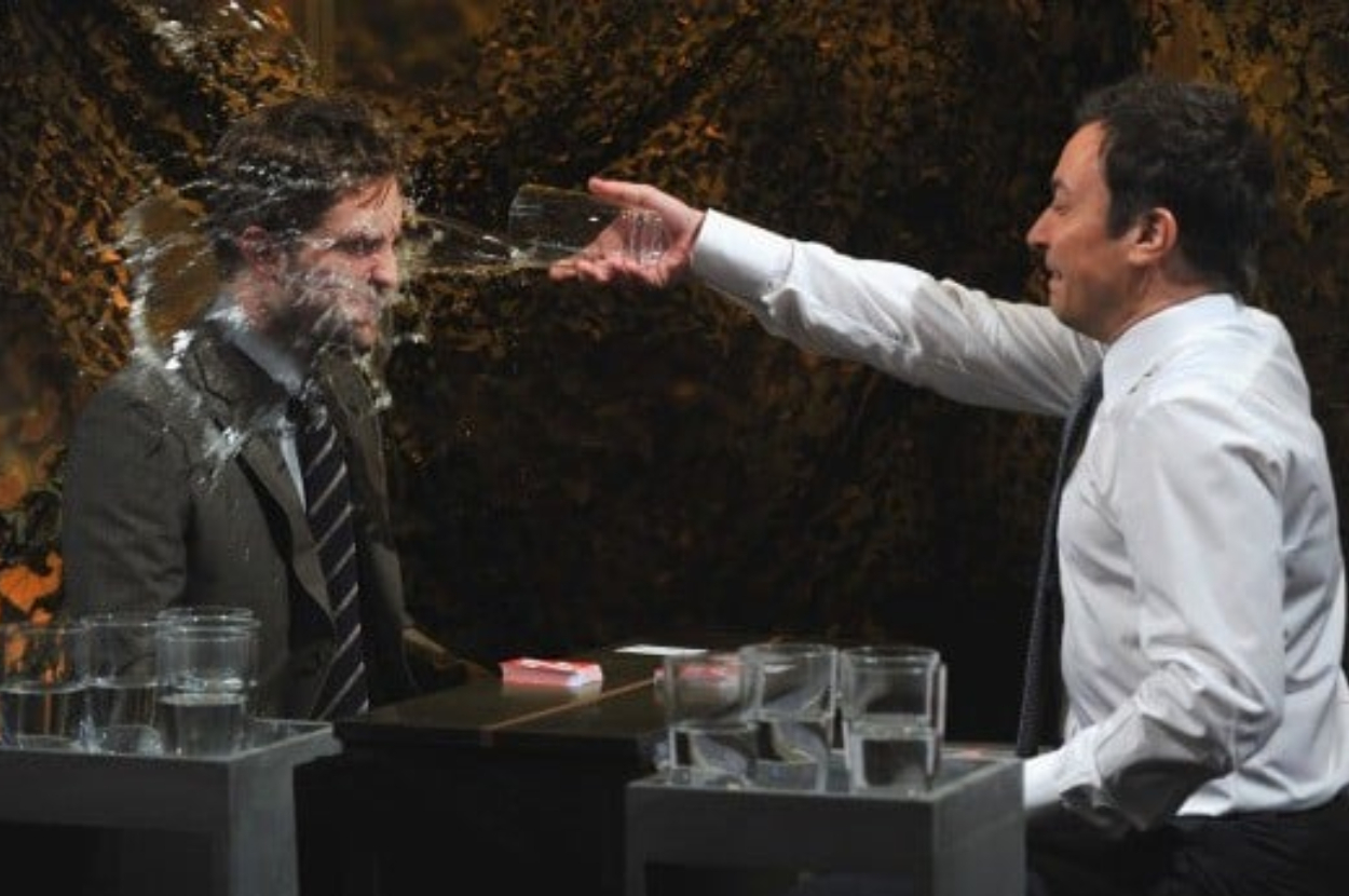Jimmy Fallon throws water on a guest during a talk show game. Both are smiling, engaged in playful activity