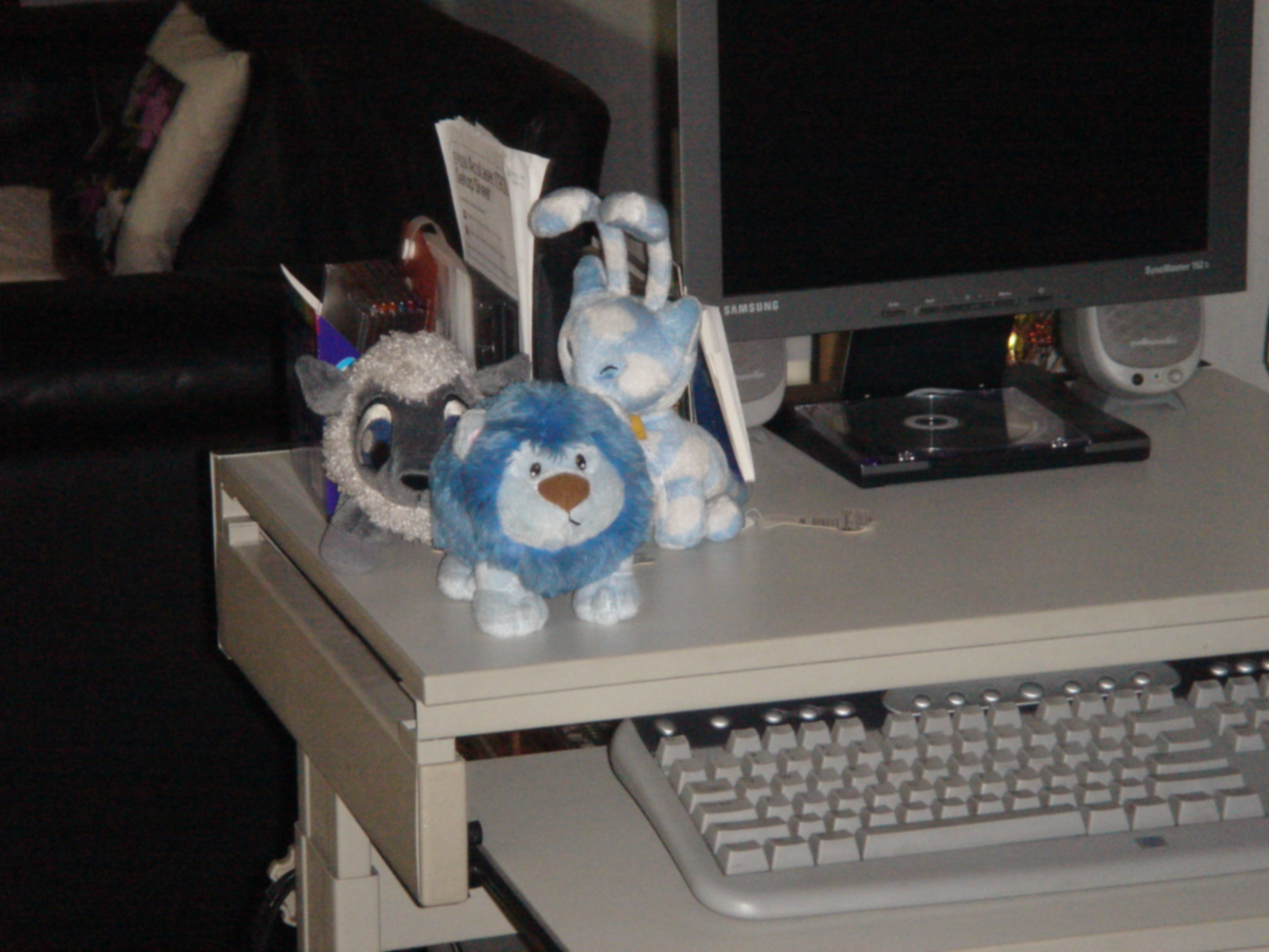 Three stuffed animal toys on a desk beside a computer keyboard and a monitor
