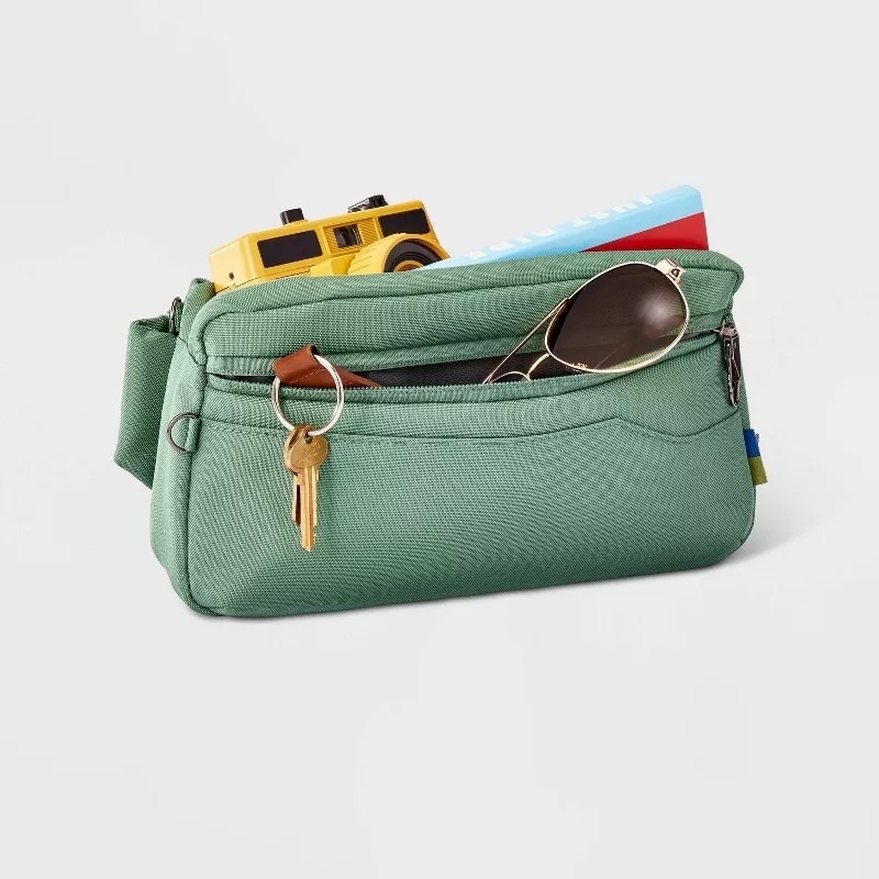 Green fanny pack with items sticking out including a camera, book, sunglasses, and keys