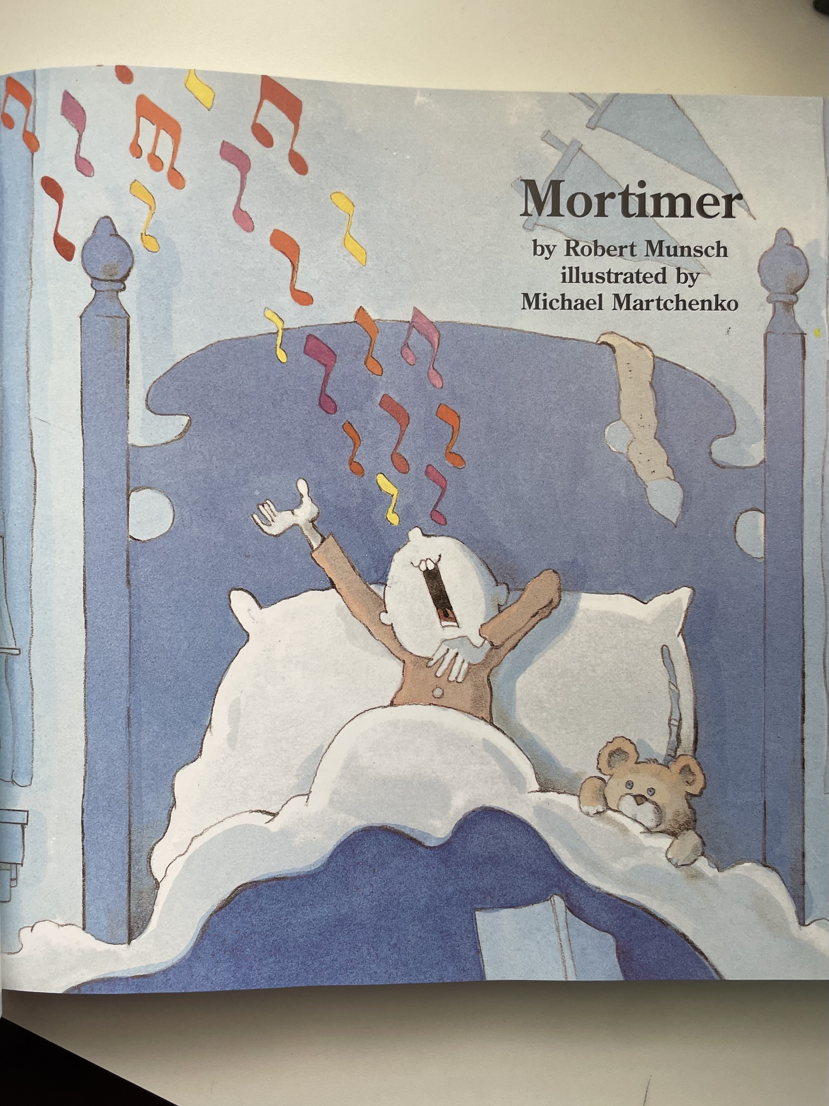 Illustration of a character, Mortimer, with a teddy bear, singing in bed from the book &quot;Mortimer&quot; by Robert Munsch