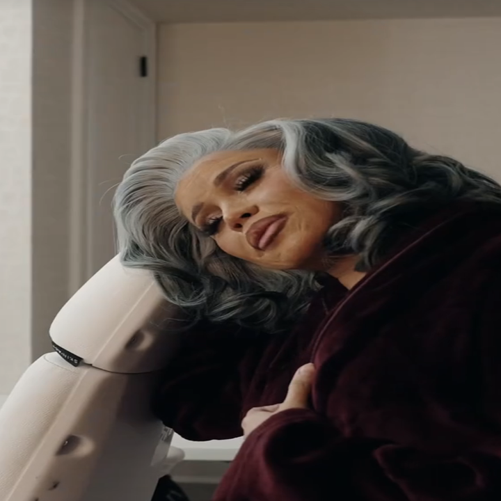 Individual with styled gray hair and makeup leaning on a chair back in a burgundy robe, giving a pensive look