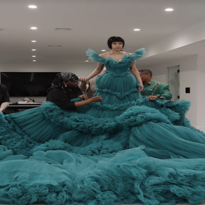 Individual in an expansive teal gown being styled by two people