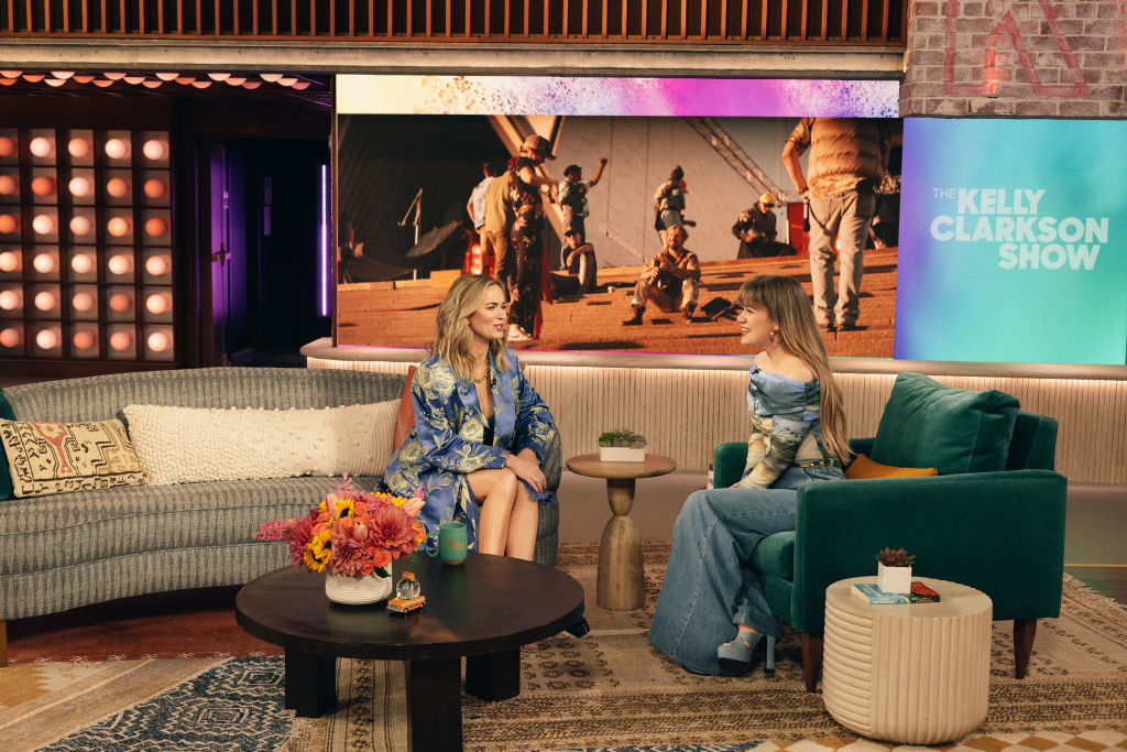 Kelly Clarkson interviews Emily Blunt on her show, both seated, with a screen showing a scene in the background