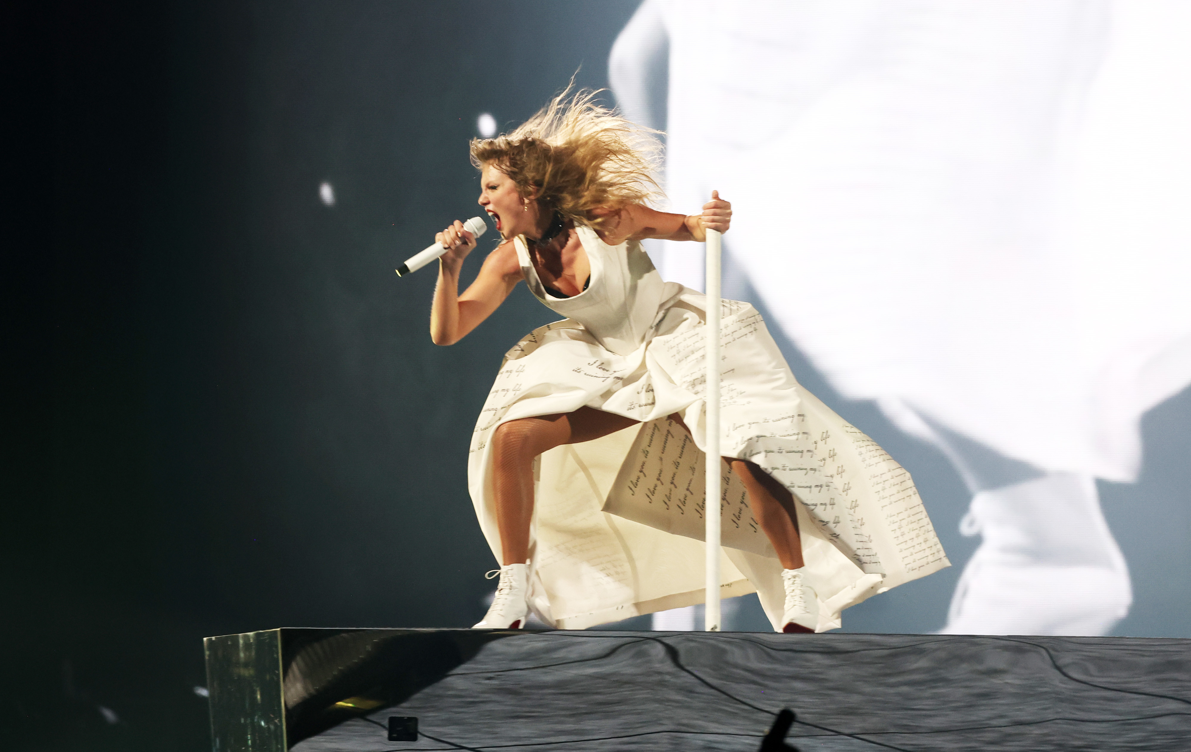 Taylor on levitating platform on stage singing into a microphone, wearing a dramatic, flowing dress with sheet music print