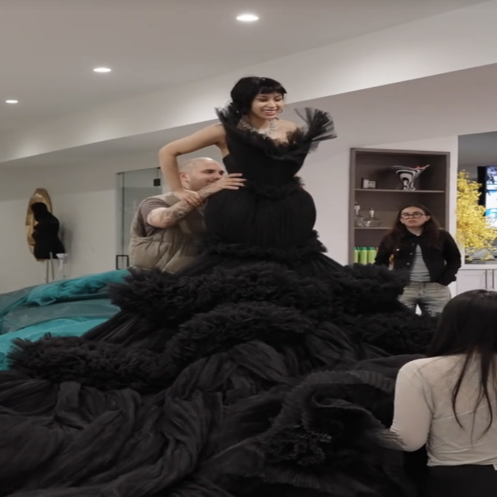 A person stands atop a voluminous black gown being arranged by others